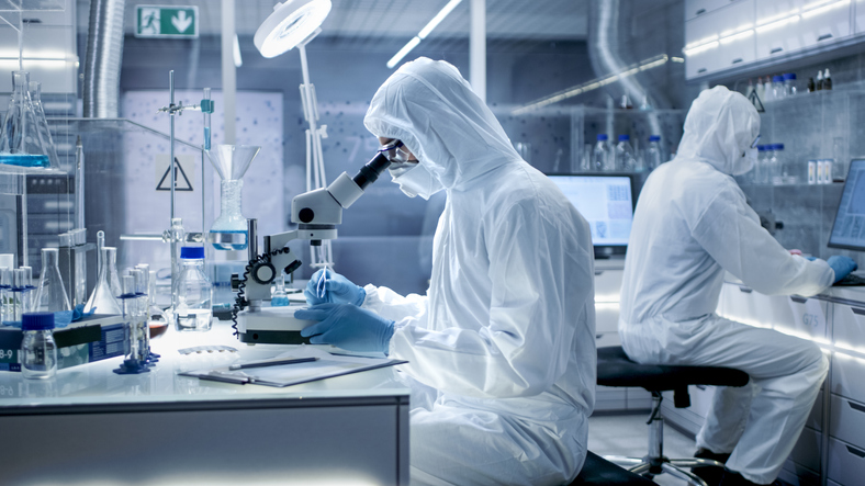 In a Secure High Level Laboratory Scientists in a Coverall Conducting a Research. Biologist Adjusts Samples in a Petri Dish with Pincers and Examines Them Under Microscope and His Colleague Analyzes Results on a Computer.