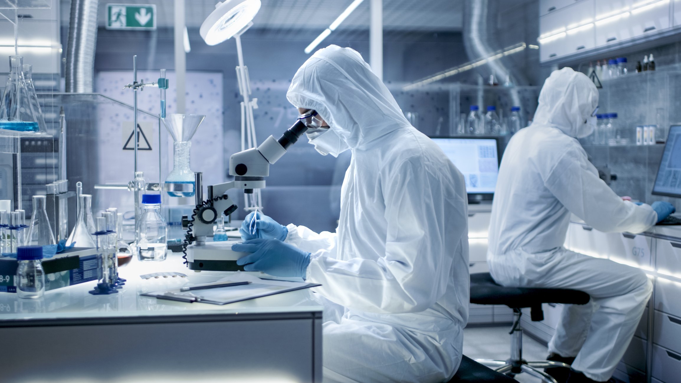 In a Secure High Level Laboratory Scientists in a Coverall Conducting a Research. Biologist Adjusts Samples in a Petri Dish with Pincers and Examines Them Under Microscope and His Colleague Analyzes Results on a Computer.